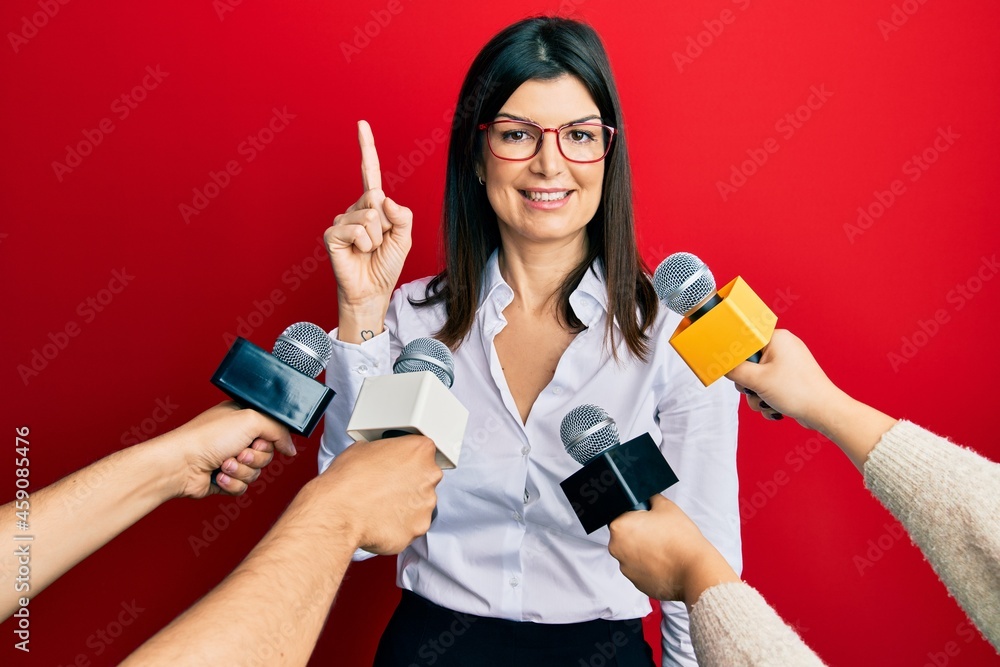 Young hispanic woman being interviewed for journalist hands with microphone smiling with an idea or question pointing finger up with happy face, number one Young hispanic woman being interviewed for journalist hands with microphone smiling with an idea or question pointing finger up with happy face, number one