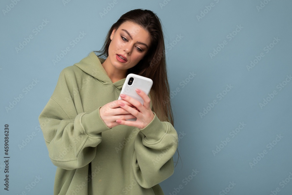 Photo of serious Beautiful young brunette woman wearing stylish green hoodie using mobile phone writing message isolated on background looking at smartphone display Photo of serious Beautiful young brunette woman wearing stylish green hoodie using mobile phone writing message isolated on background looking at smartphone display