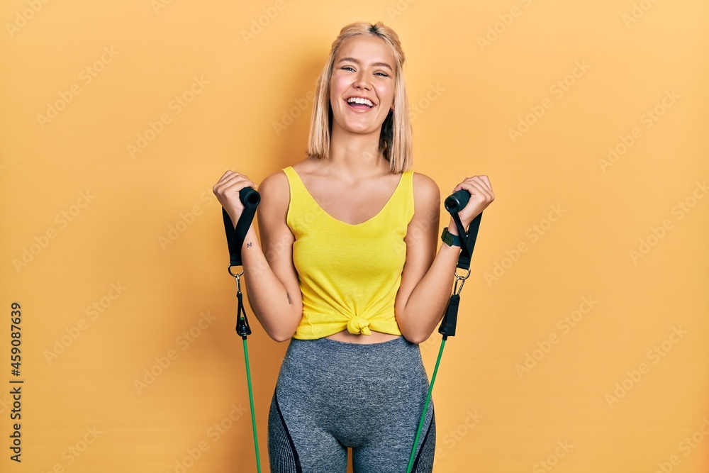 Beautiful blonde woman training arm resistance with elastic arm bands smiling and laughing hard out loud because funny crazy joke.