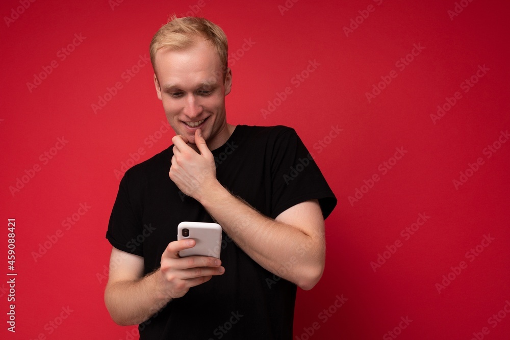 Photo shot of handsome positive good looking young man wearing casual stylish outfit poising isolated on background with empty space holding in hand and using mobile phone messaging sms looking at Photo shot of handsome positive good looking young man wearing casual stylish outfit poising isolated on background with empty space holding in hand and using mobile phone messaging sms looking at