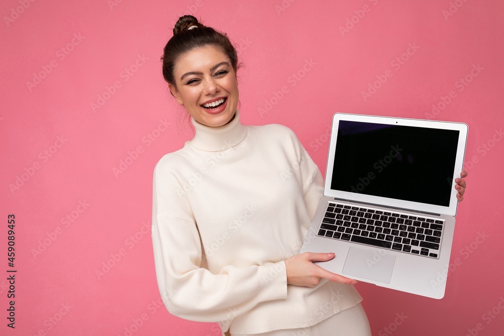 Photo of beautiful smiling and laughung brunette young lady with gathered dark hair wearing white sweater holding computer laptop and looking at camera with close eyes and open mouth isolated on pink Photo of beautiful smiling and laughung brunette young lady with gathered dark hair wearing white sweater holding computer laptop and looking at camera with close eyes and open mouth isolated on pink