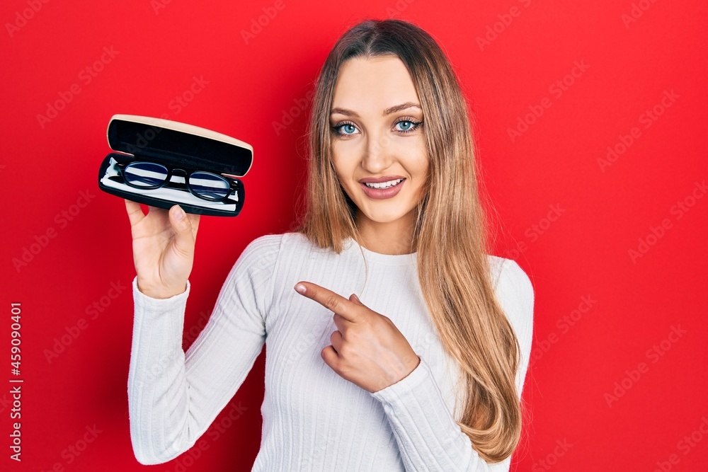 Young blonde girl holding glasses smiling happy pointing with hand and finger Young blonde girl holding glasses smiling happy pointing with hand and finger