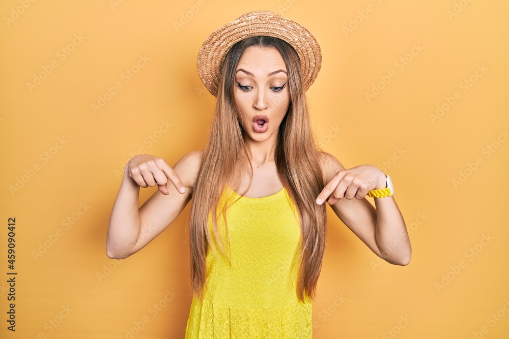 Young blonde girl wearing summer hat pointing down with fingers showing advertisement, surprised face and open mouth Young blonde girl wearing summer hat pointing down with fingers showing advertisement, surprised face and open mouth
