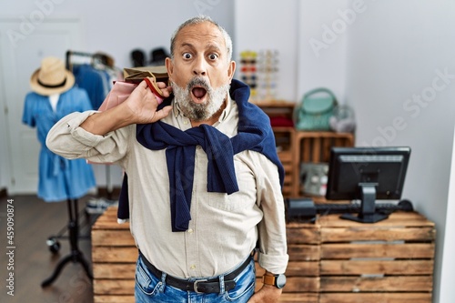 Handsome senior man holding shopping bags at boutique shop in shock face, looking skeptical and sarcastic, surprised with open mouth