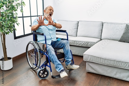 Handsome senior man sitting on wheelchair at the living room afraid and terrified with fear expression stop gesture with hands, shouting in shock. panic concept.