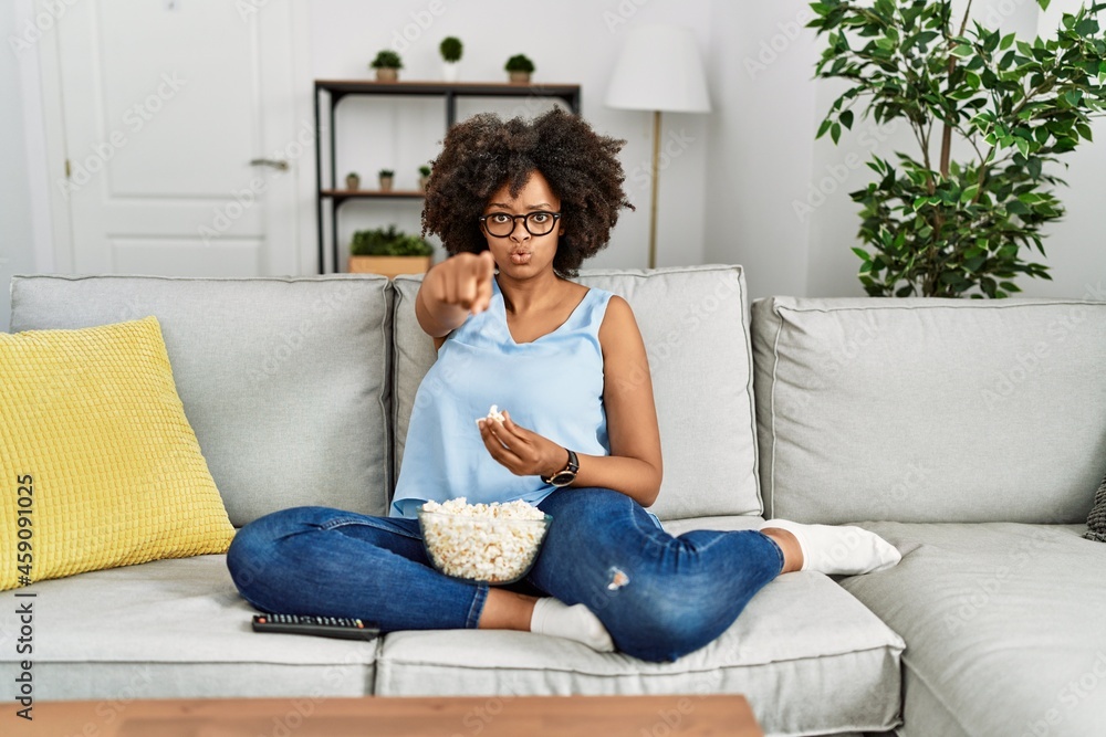 African american woman with afro hair sitting on the sofa eating popcorn at home pointing with finger to the camera and to you, confident gesture looking serious African american woman with afro hair sitting on the sofa eating popcorn at home pointing with finger to the camera and to you, confident gesture looking serious