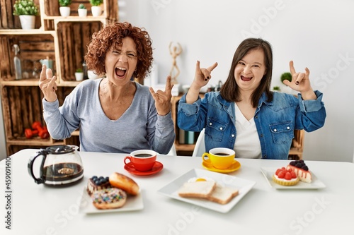 Family of mother and down syndrome daughter sitting at home eating breakfast shouting with crazy expression doing rock symbol with hands up. music star. heavy concept.