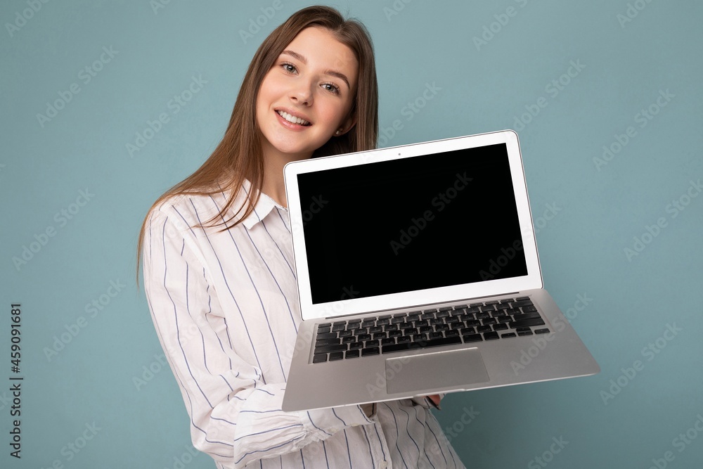 Close-up portrait of Beautiful smiling young woman holding netbook computer looking at camera wearing white shirt isolated on blue background Close-up portrait of Beautiful smiling young woman holding netbook computer looking at camera wearing white shirt isolated on blue background