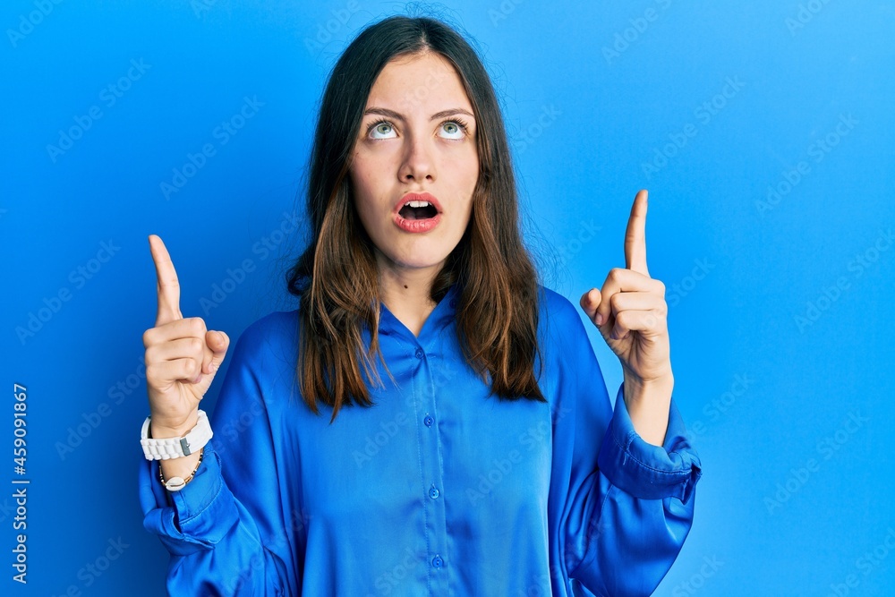 Young brunette woman wearing casual blue shirt amazed and surprised looking up and pointing with fingers and raised arms. Young brunette woman wearing casual blue shirt amazed and surprised looking up and pointing with fingers and raised arms.