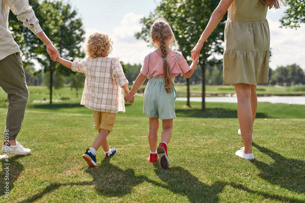 Cropped shot of happy young family holding hands, walking together outside in green summer park