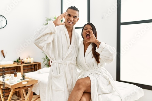 Young latin couple wearing towel standing at beauty center shouting and screaming loud to side with hand on mouth. communication concept.