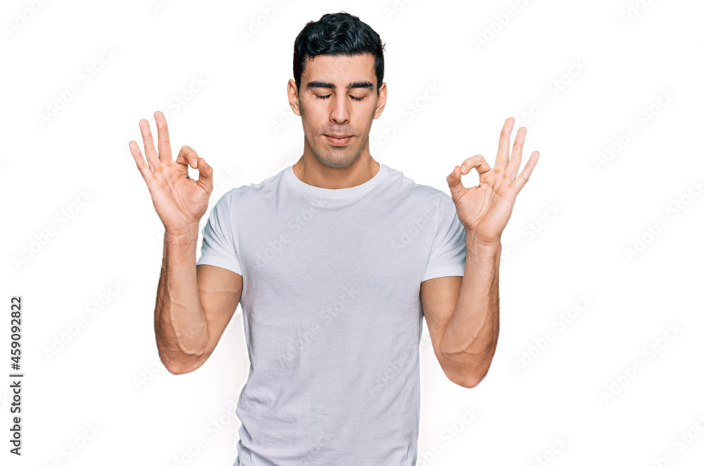 Handsome hispanic man wearing casual white t shirt relax and smiling with eyes closed doing meditation gesture with fingers. yoga concept. Handsome hispanic man wearing casual white t shirt relax and smiling with eyes closed doing meditation gesture with fingers. yoga concept.