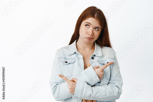 Confused young redhead woman pointing sideways and looking up, thinking what to choose, picking from two variants, white background