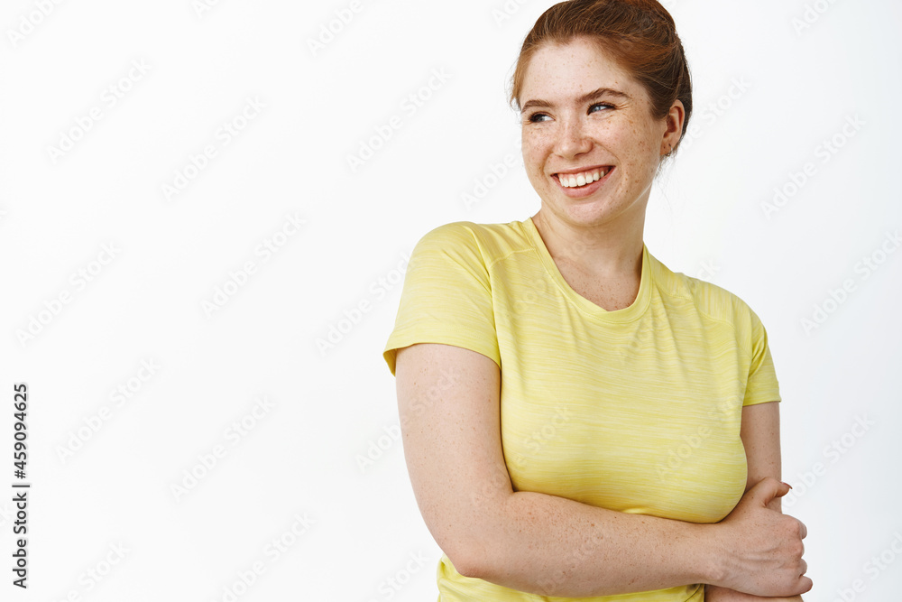 Close up of carefree redhead woman in fitness t-shirt, looking away and smiling, workout in gym, standing over white background Close up of carefree redhead woman in fitness t-shirt, looking away and smiling, workout in gym, standing over white background