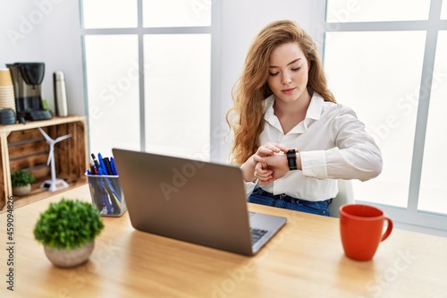 Young caucasian woman working at the office using computer laptop checking the time on wrist watch, relaxed and confident