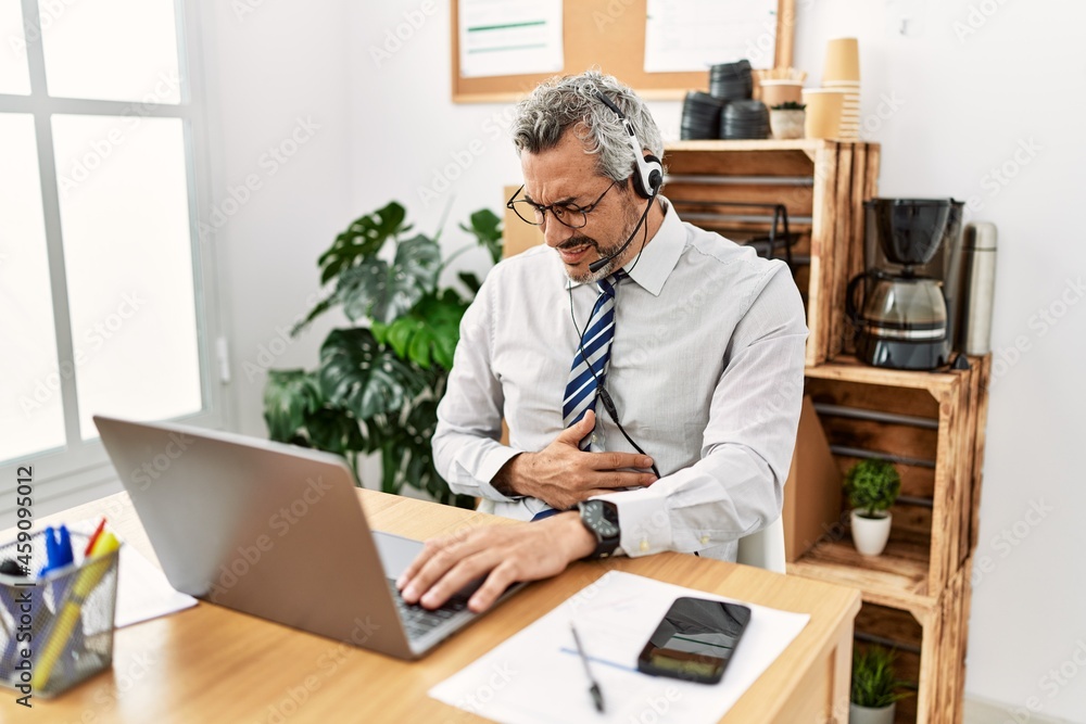 Middle age hispanic business man working at the office wearing operator headset with hand on stomach because nausea, painful disease feeling unwell. ache concept. Middle age hispanic business man working at the office wearing operator headset with hand on stomach because nausea, painful disease feeling unwell. ache concept.