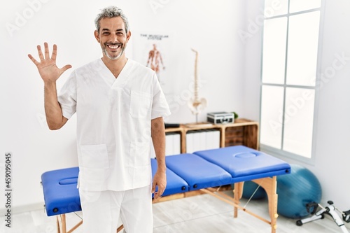 Middle age hispanic therapist man working at pain recovery clinic showing and pointing up with fingers number five while smiling confident and happy.