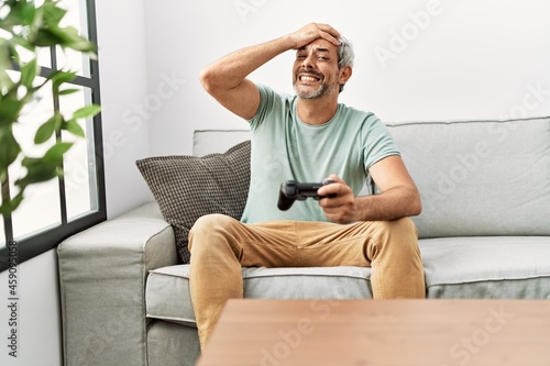 Middle age hispanic man playing video game sitting on the sofa stressed and frustrated with hand on head, surprised and angry face