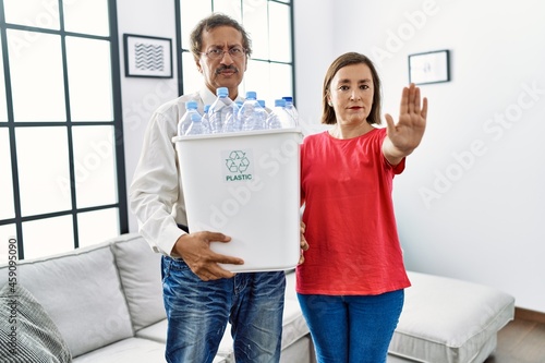 Middle age interracial couple holding recycling bin with plastic bottles at home with open hand doing stop sign with serious and confident expression, defense gesture