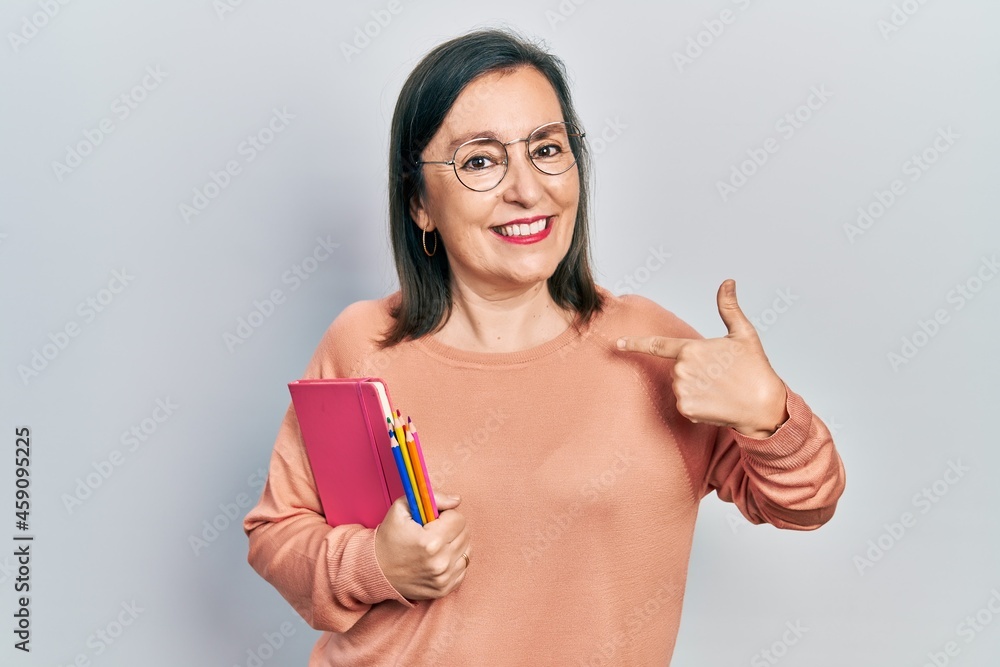 Middle age hispanic woman holding book and color pencils pointing finger to one self smiling happy and proud Middle age hispanic woman holding book and color pencils pointing finger to one self smiling happy and proud