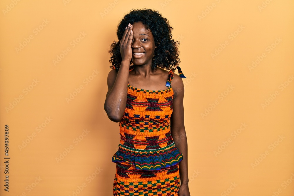 Young african american woman wearing traditional african clothes covering one eye with hand, confident smile on face and surprise emotion. Young african american woman wearing traditional african clothes covering one eye with hand, confident smile on face and surprise emotion.