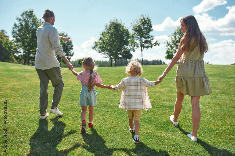 Back view of happy family with little kids, boy and girl holding hands together and walking on green grass field in the park on a summer day