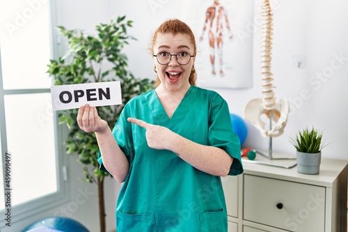 Young redhead physiotherapist woman working at pain recovery clinic holding open banner smiling happy pointing with hand and finger