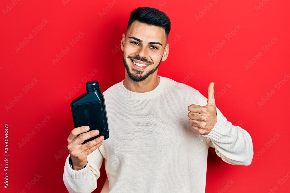Young hispanic man with beard holding motor oil bottle smiling happy and positive, thumb up doing excellent and approval sign Young hispanic man with beard holding motor oil bottle smiling happy and positive, thumb up doing excellent and approval sign