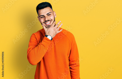 Young hispanic man with beard wearing casual orange sweater looking confident at the camera smiling with crossed arms and hand raised on chin. thinking positive.