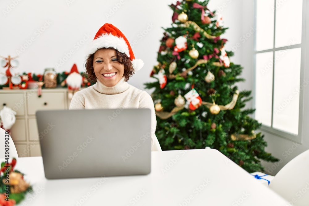 Middle age hispanic woman using laptop sitting by christmas tree looking positive and happy standing and smiling with a confident smile showing teeth Middle age hispanic woman using laptop sitting by christmas tree looking positive and happy standing and smiling with a confident smile showing teeth
