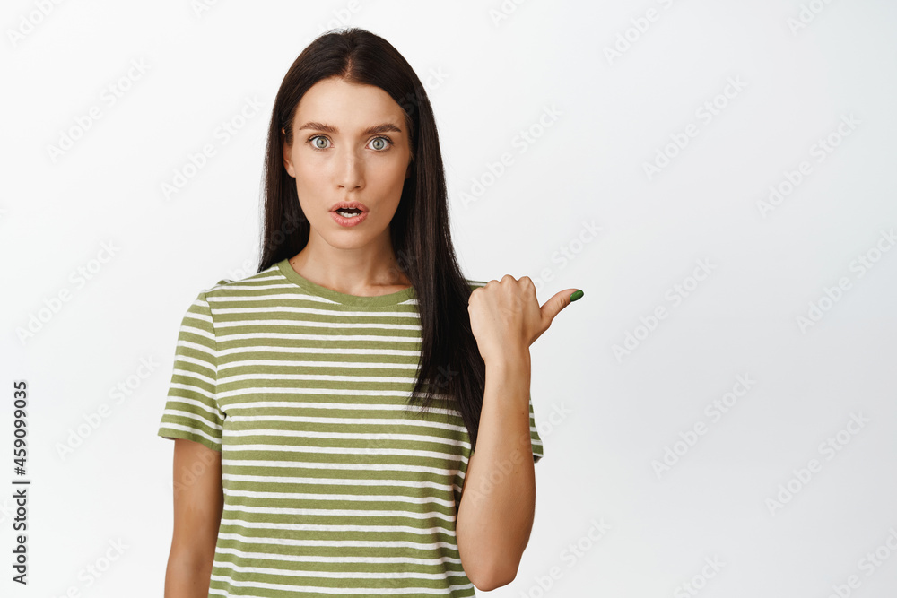 Image of shocked brunette woman pointing right, looking amazed at camera, showing smth amazing, standing over white background Image of shocked brunette woman pointing right, looking amazed at camera, showing smth amazing, standing over white background