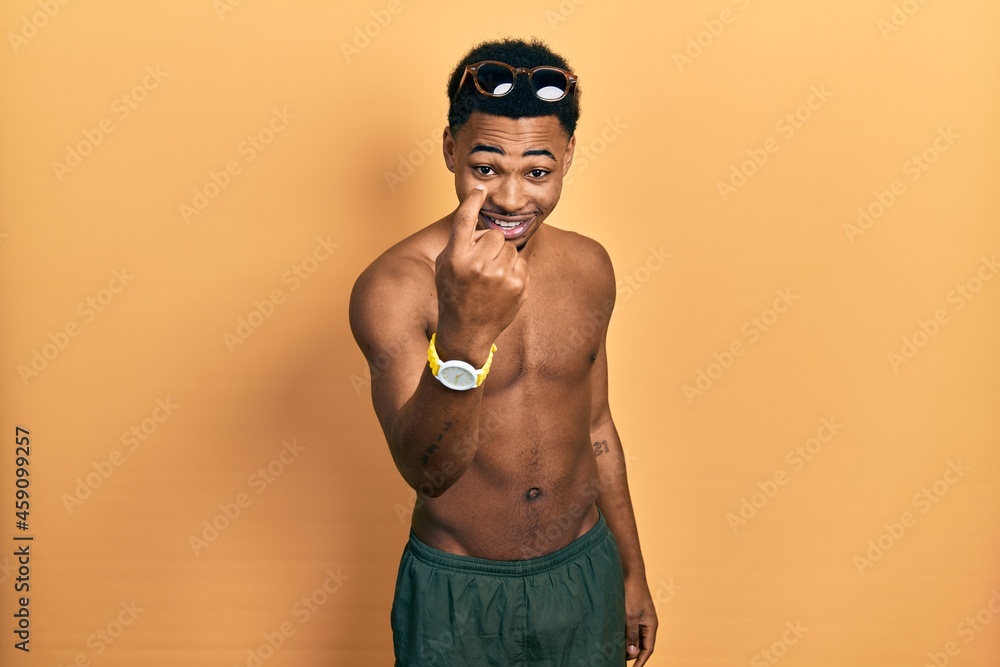 Young african american man wearing swimwear and swimmer glasses beckoning come here gesture with hand inviting welcoming happy and smiling