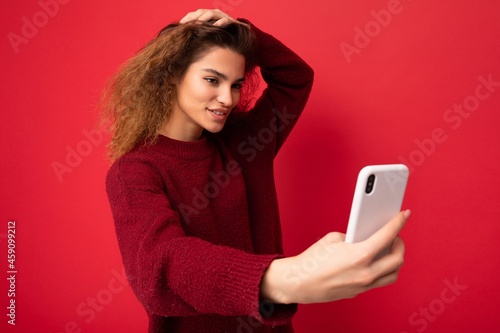 Shot of sexy smiling Beautiful young woman with curly hair wearing dark red sweater isolated on red background wall holding and using smart phone looking at telephone screen and taking selfie photo