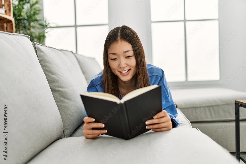 Young chinese girl reading book lying on the sofa at home. Young chinese girl reading book lying on the sofa at home.