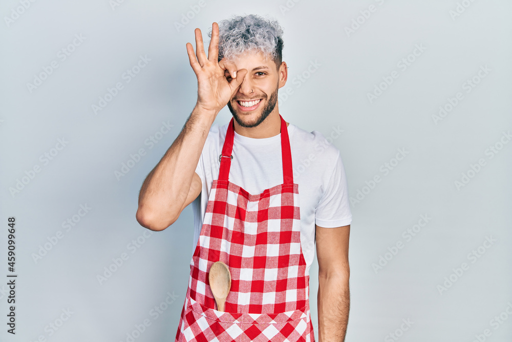 Young hispanic man with modern dyed hair wearing apron doing ok gesture with hand smiling, eye looking through fingers with happy face. Young hispanic man with modern dyed hair wearing apron doing ok gesture with hand smiling, eye looking through fingers with happy face.