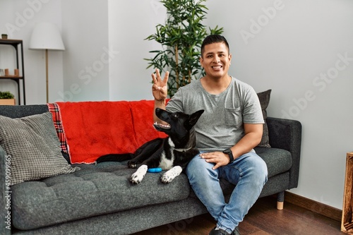 Young latin man and dog sitting on the sofa at home showing and pointing up with fingers number three while smiling confident and happy. © Krakenimages.com