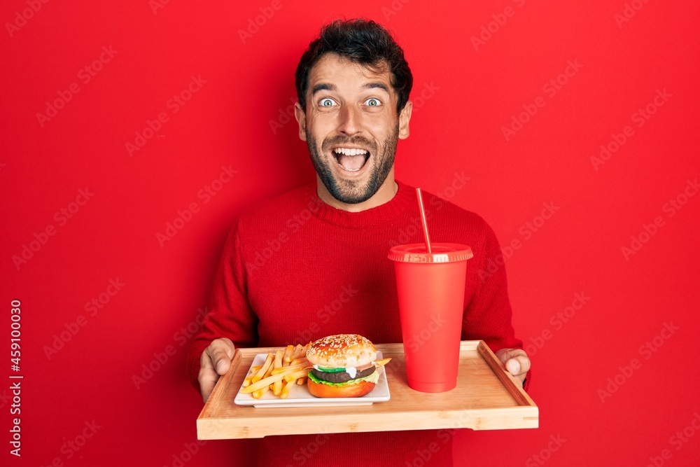 Handsome man with beard eating a tasty classic burger with fries and soda celebrating crazy and amazed for success with open eyes screaming excited. Handsome man with beard eating a tasty classic burger with fries and soda celebrating crazy and amazed for success with open eyes screaming excited.