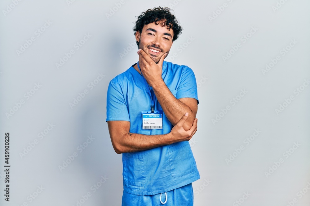 Young hispanic man wearing blue male nurse uniform looking confident at the camera smiling with crossed arms and hand raised on chin. thinking positive.
