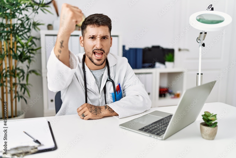 Young doctor working at the clinic using computer laptop angry and mad raising fist frustrated and furious while shouting with anger. rage and aggressive concept. Young doctor working at the clinic using computer laptop angry and mad raising fist frustrated and furious while shouting with anger. rage and aggressive concept.