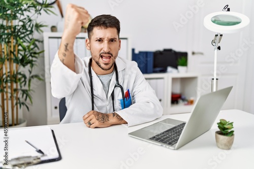 Young doctor working at the clinic using computer laptop angry and mad raising fist frustrated and furious while shouting with anger. rage and aggressive concept.