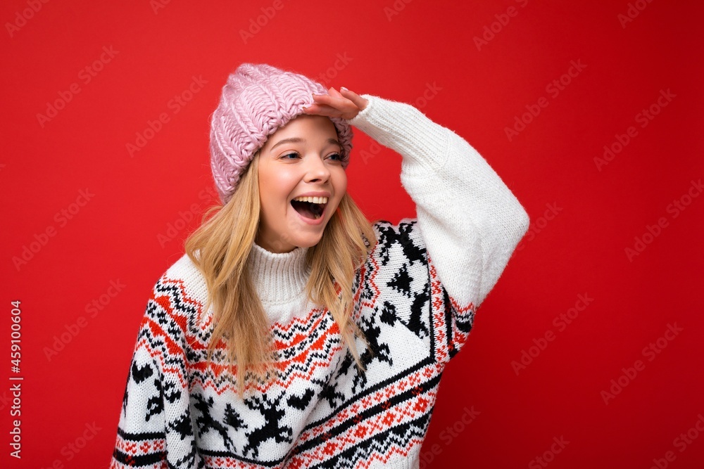 Photo of young positive happy beautiful blonde woman with sincere emotions wearing pink knitted hat and winter pullover isolated over red background with empty space and looking to the side with hand Photo of young positive happy beautiful blonde woman with sincere emotions wearing pink knitted hat and winter pullover isolated over red background with empty space and looking to the side with hand
