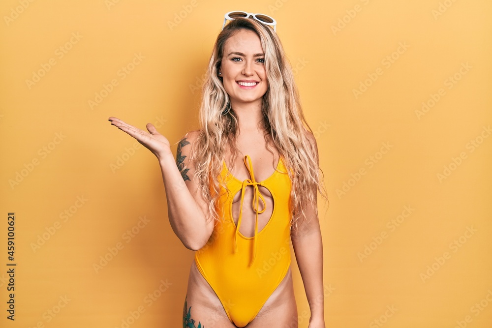 Beautiful young blonde woman wearing swimsuit and sunglasses smiling cheerful presenting and pointing with palm of hand looking at the camera.