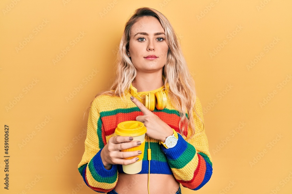 Beautiful young blonde woman drinking cup of coffee wearing headphones pointing with hand finger to the side showing advertisement, serious and calm face Beautiful young blonde woman drinking cup of coffee wearing headphones pointing with hand finger to the side showing advertisement, serious and calm face