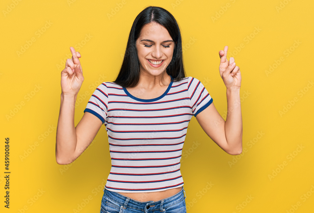 Young hispanic girl wearing casual striped t shirt gesturing finger crossed smiling with hope and eyes closed. luck and superstitious concept. Young hispanic girl wearing casual striped t shirt gesturing finger crossed smiling with hope and eyes closed. luck and superstitious concept.