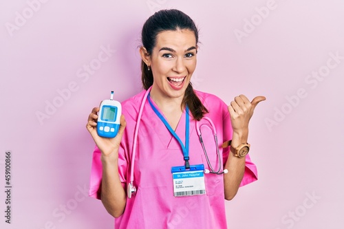 Young brunette woman wearing doctor uniform holding glucometer pointing thumb up to the side smiling happy with open mouth