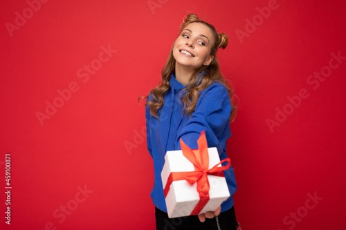 Fascinating happy smiling amazing young blonde woman isolated over red background wall wearing blue stylish hoodie holding gift box and looking at camera