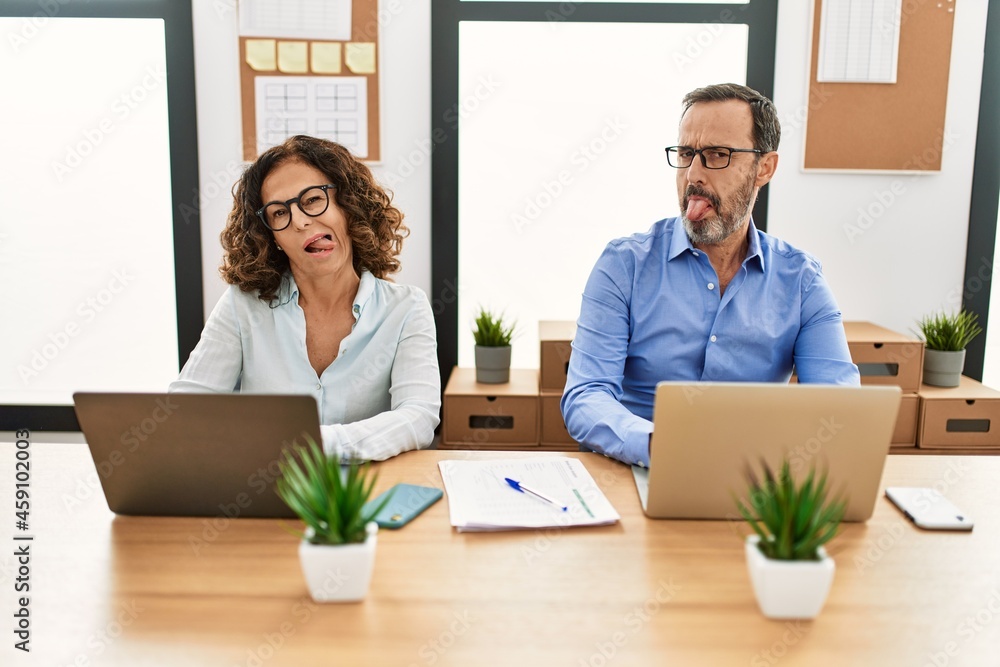 Middle age hispanic woman and man sitting with laptop at the office sticking tongue out happy with funny expression. emotion concept.