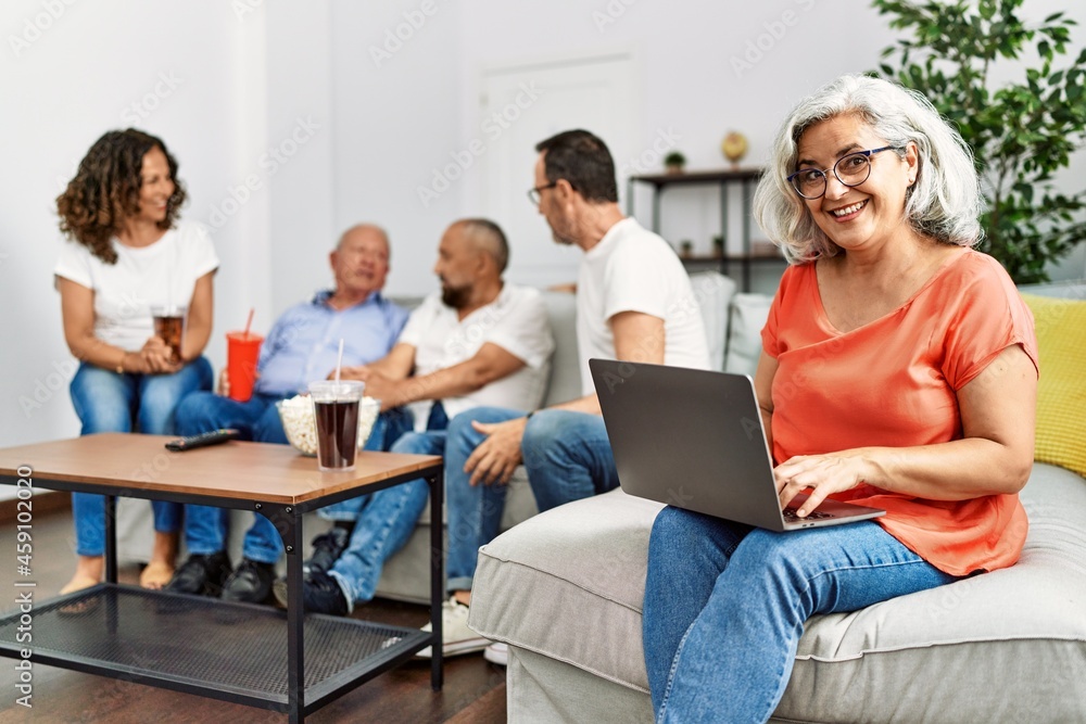 Fototapeta premium Group of middle age friends sitting on the sofa speaking. Woman smiling happy using laptop at home.
