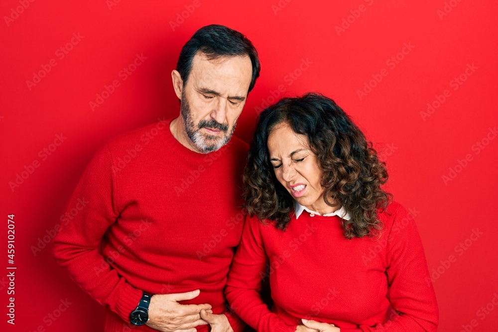 Middle age couple of hispanic woman and man hugging and standing together with hand on stomach because indigestion, painful illness feeling unwell. ache concept. Middle age couple of hispanic woman and man hugging and standing together with hand on stomach because indigestion, painful illness feeling unwell. ache concept.