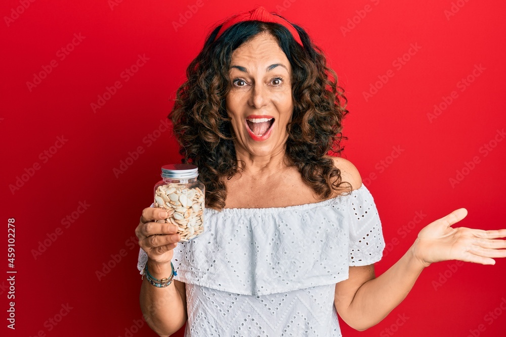Middle age hispanic woman holding jar with pumpkin seeds celebrating achievement with happy smile and winner expression with raised hand Middle age hispanic woman holding jar with pumpkin seeds celebrating achievement with happy smile and winner expression with raised hand